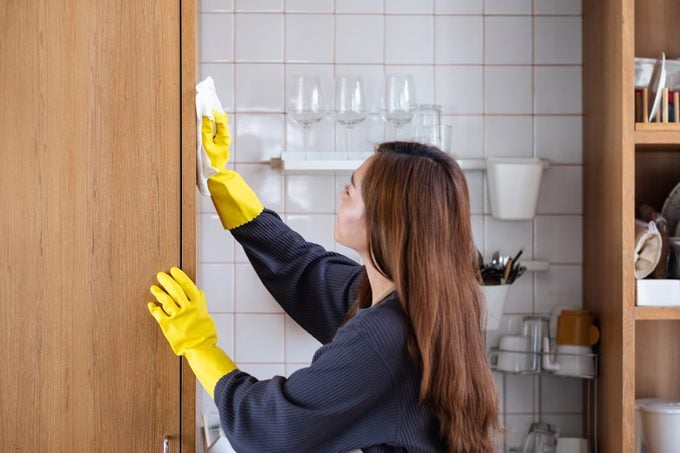 Una mujer con guante protector, limpiando un armario de madera en la cocina de casa